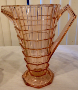 Depression class, c. 1926. Pink depression glass pitcher on an octagonal foot; body tapers from top to bottom in a series of eight rings, eight sections in each ring. Angular spout and angular handle. Inside wall of pitcher is rounded.